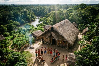 Vista aérea de uma densa floresta preservada no Amazonas com luz solar entre as árvores. Representantes de comunidade tradicional na Amazônia conversando em frente a uma maloca de madeira.